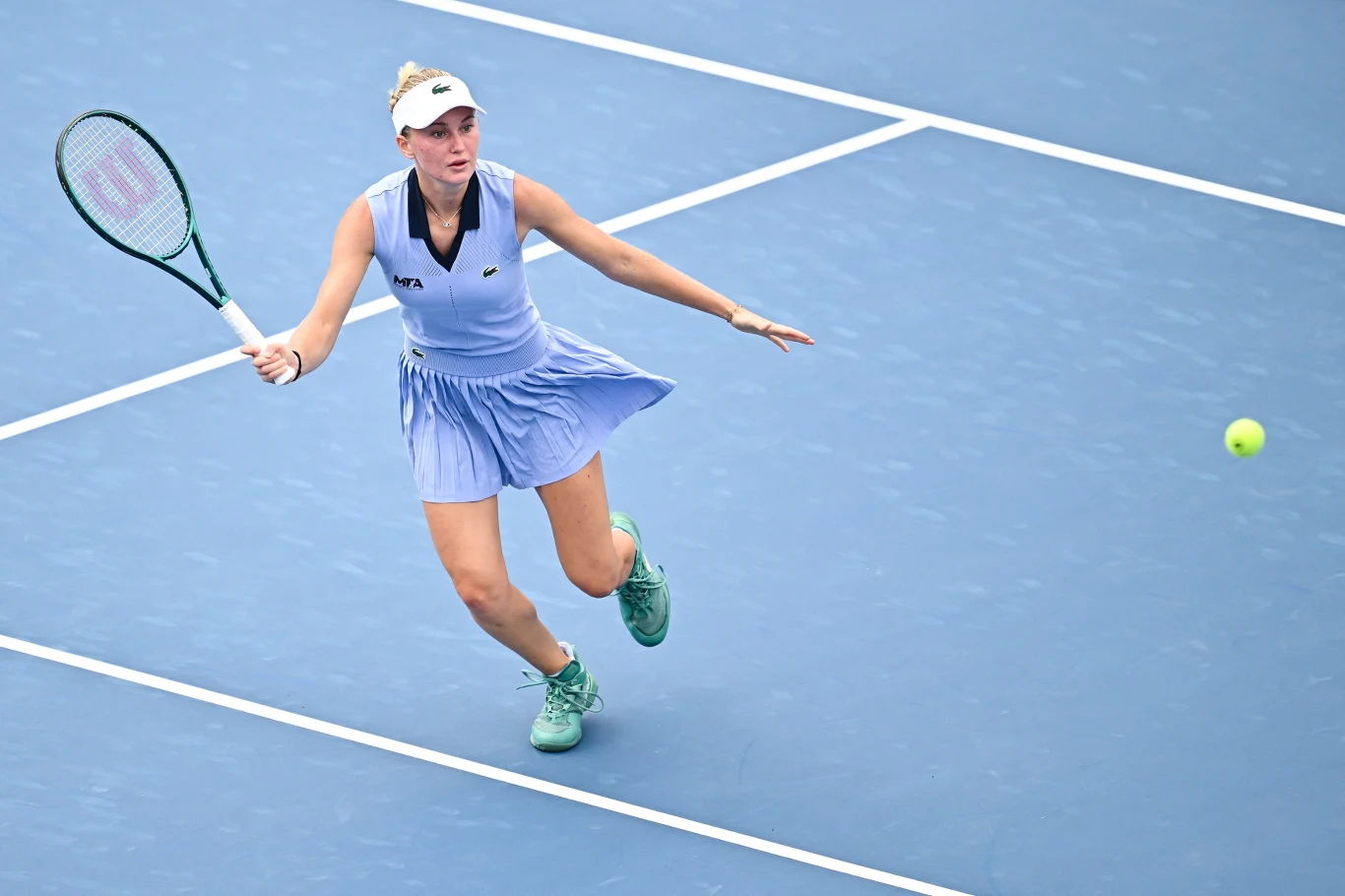 Tenisistka w niebieskiej sukience podczas gry na kortach twardych, biegnąca w kierunku piłki, skoncentrowana na odbiciu, w dynamicznej pozie.