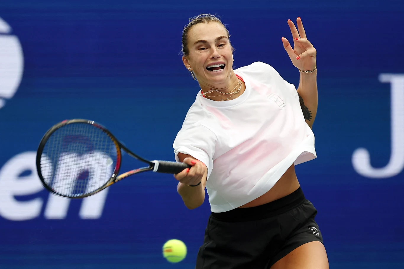 Kobieta w sportowym stroju dynamicznie odbija piłkę tenisową rakietą na korcie podczas meczu tenisowego, skupiona na grze, z widocznym logotypem turnieju w tle.