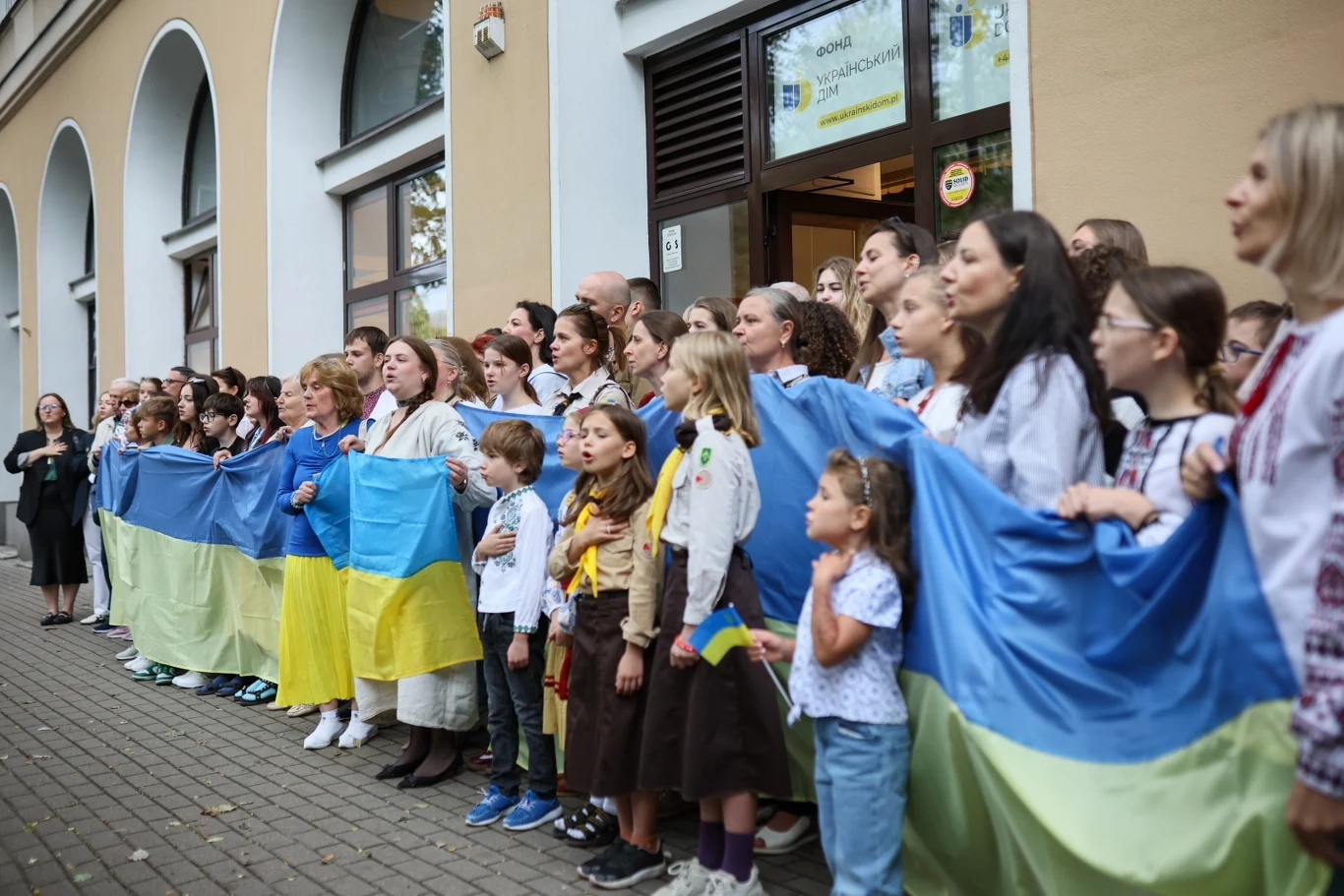 Grupa osób, głównie kobiet i dzieci, stoi blisko siebie trzymając dużą flagę Ukrainy na tle budynku z łukowatymi oknami. Uczestnicy mają na sobie tradycyjne stroje lub elementy w barwach narodowych, panuje atmosfera wspólnoty i jedności.