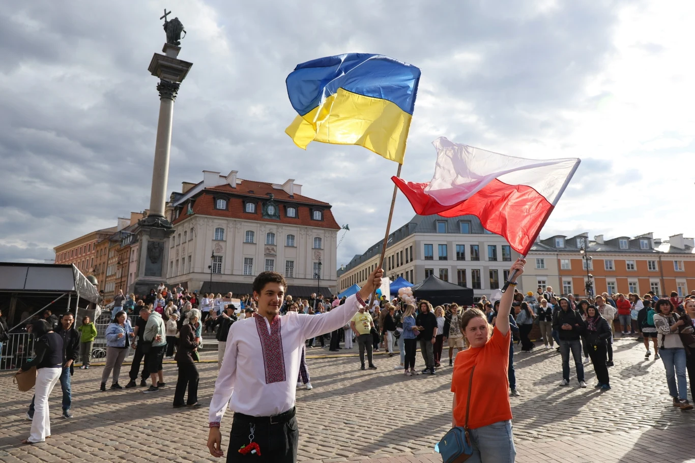 Dwie osoby trzymające flagi Ukrainy i Polski na tle Kolumny Zygmunta i starego miasta, wokół zgromadzeni ludzie, wydarzenie odbywa się na otwartym placu.