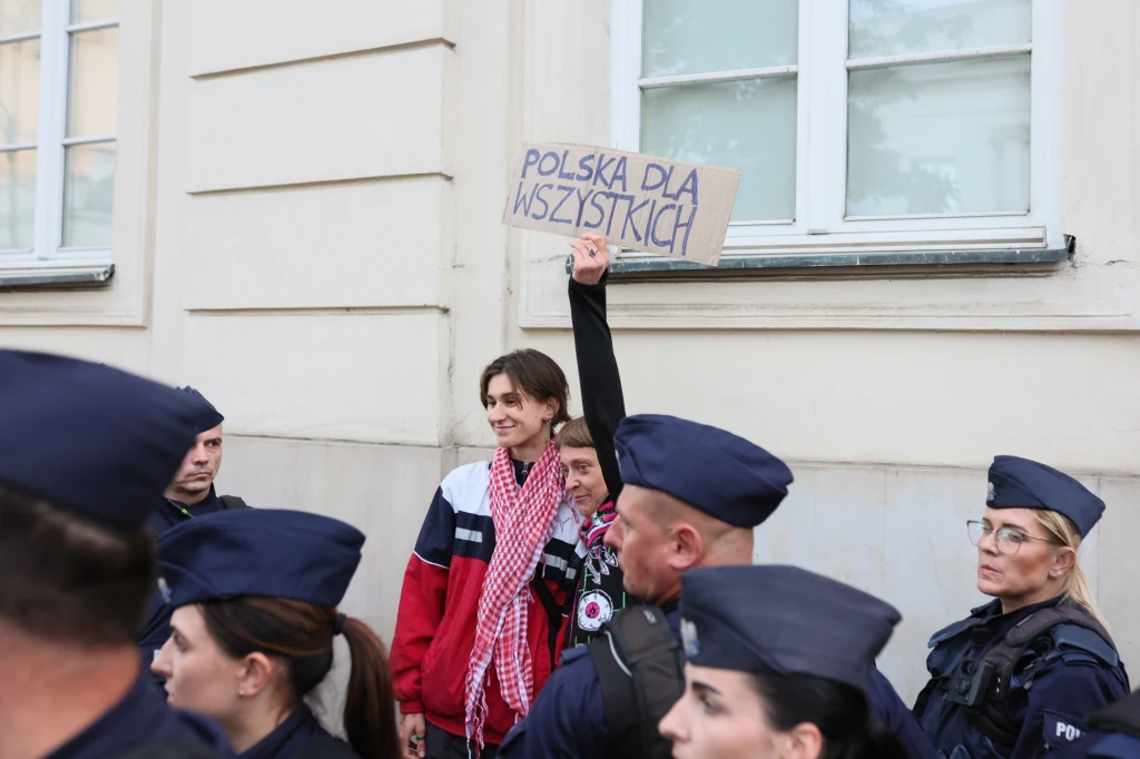 Grupa policjantów pilnujących porządku wokół osoby stojącej pod budynkiem, trzymającej kartonowy transparent z napisem 'Polska dla wszystkich', wyrażający postulat równości.