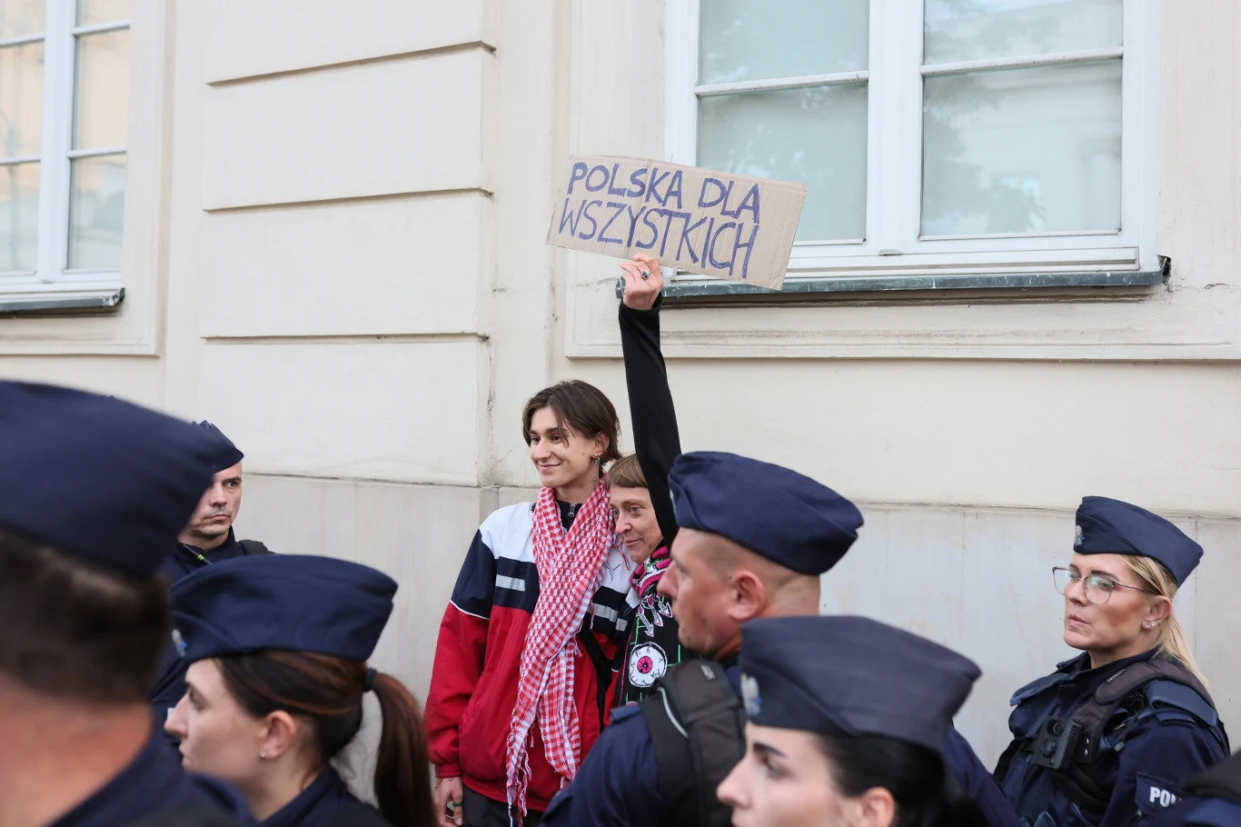 Grupa policjantów pilnujących porządku wokół osoby stojącej pod budynkiem, trzymającej kartonowy transparent z napisem 'Polska dla wszystkich', wyrażający postulat równości.
