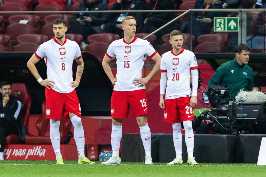 Jakub Moder, Adam Buksa, Sebastian Szymański Trzech piłkarzy w biało-czerwonych strojach reprezentacji Polski stoi na murawie stadionu, za nimi widoczna jest ławka rezerwowych oraz osoby z obsługi technicznej, na trybunach nieliczni widzowie