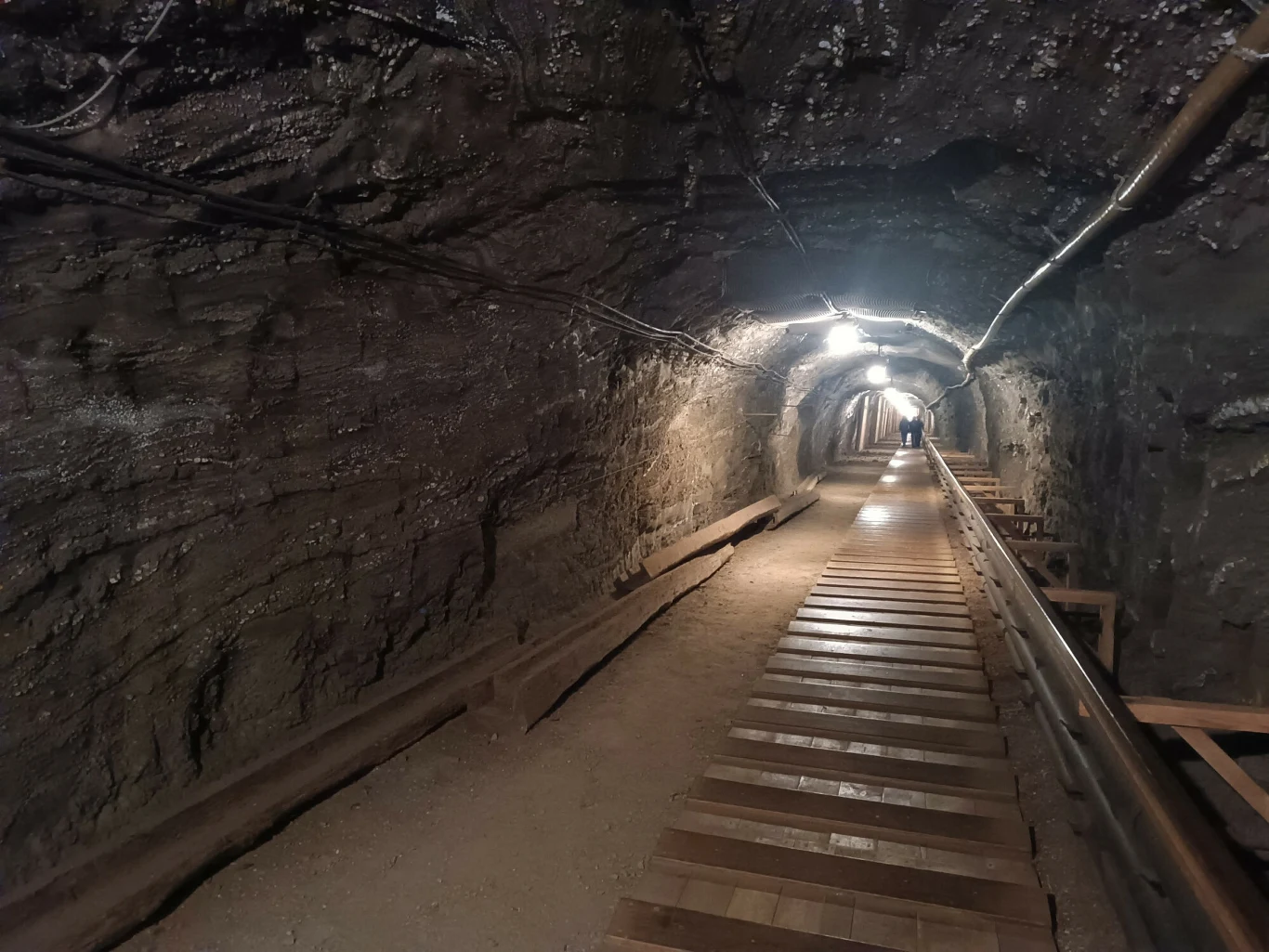 Wnętrze Kopalni Soli Bochnia Drewniana kładka prowadzi przez oświetlony tunel wykuty w skale, po bokach widać metalowe rury biegnące wzdłuż ścian, sufit pokryty jest wilgotną skałą.