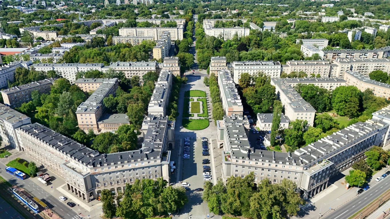 Nowa Huta. Widok na al. Róż i Plac Centralny im. Ronalda Reagana Zabudowa miejska o charakterystycznym układzie symetrycznym z centralnym placem zielonym, otoczona blokami mieszkalnymi i szerokimi alejami, całość otulona dużą ilością drzew.