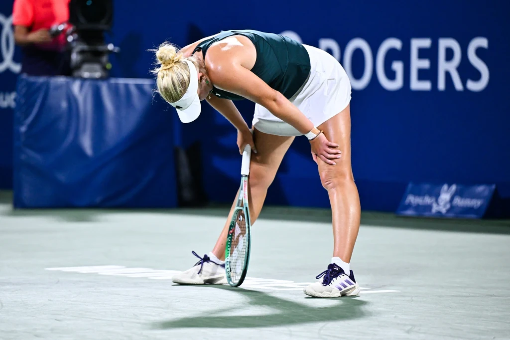 Clara Tauson Tenisistka w białej spódniczce i zielonym topie opiera się na rakiecie podczas przerwy na korcie tenisowym, pochylona do przodu z widocznym zmęczeniem.