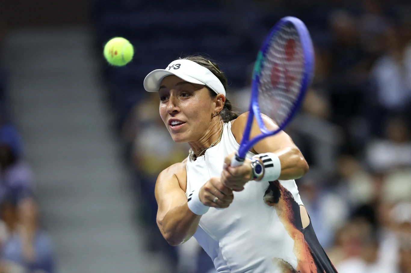 Tenisistka w białej czapce i stroju sportowym wykonuje dwuręczny backhand, skupiona na odbijaniu zbliżającej się piłki tenisowej podczas meczu na korcie.