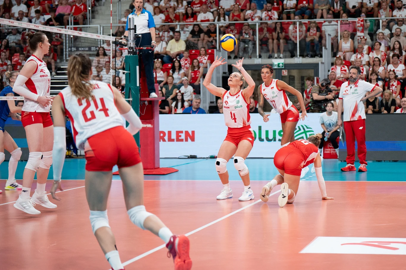 Drużyna żeńska podczas meczu siatkówki halowej, zawodniczki w biało-czerwonych strojach koncentrują się na piłce przelatującej nad siatką, a widownia pełna kibiców żywiołowo dopinguje z trybun.