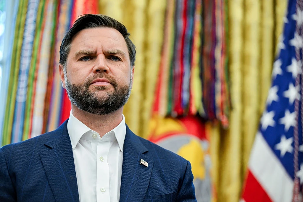 Wiceprezydent USA JD Vance Mężczyzna o poważnym wyrazie twarzy w granatowej marynarce i białej koszuli stojący na tle żółtej zasłony oraz flag, w tym flagi Stanów Zjednoczonych.