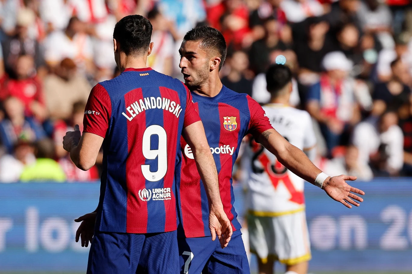 Dwóch piłkarzy drużyny FC Barcelona w charakterystycznych granatowo-bordowych strojach dyskutuje na boisku podczas meczu piłkarskiego, tłum oraz zawodnicy drużyny przeciwnej w tle.