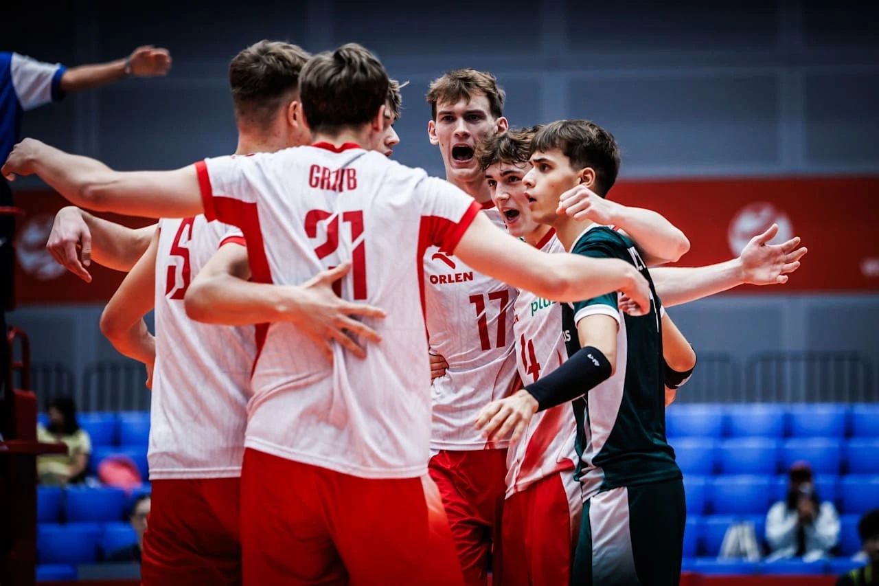 Grupa młodych siatkarzy ubranych w biało-czerwone stroje świętuje punkt podczas meczu, w tle widać trybuny oraz zawodników przeciwnika.