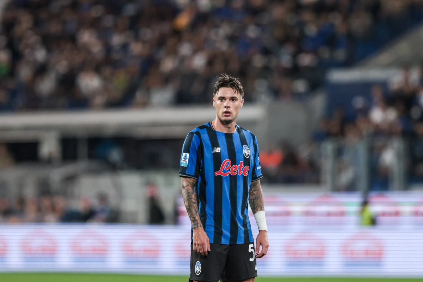 Nicola Zalewski niedawno przeszedł do Atalanty z Interu Mediolan Piłkarz w czarno-niebieskiej koszulce z widocznymi tatuażami na przedramionach stoi na stadionie, wokół niego rozmazane sylwetki kibiców i trybuny.