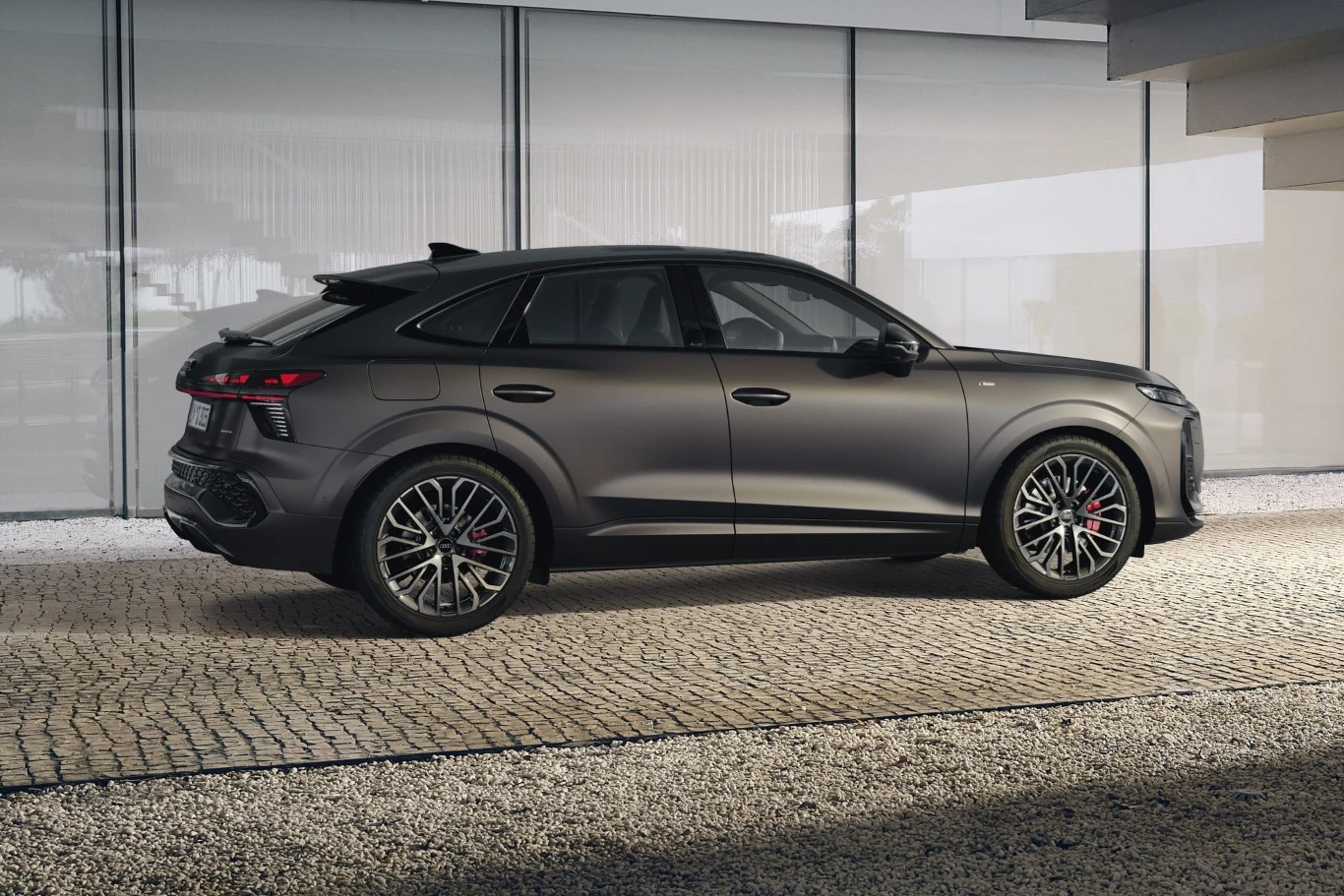 Luksusowy, sportowy SUV w kolorze matowej szarości zaparkowany na brukowanej nawierzchni, widok z boku na tle nowoczesnego, przeszklonego budynku; auto wyróżnia się dużymi felgami i aerodynamiczną sylwetką.