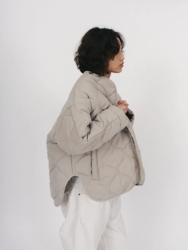 Kurtka oversize pique light beige Kobieta o ciemnych, kręconych włosach ubrana w jasną, pikowaną kurtkę i białe spodnie pozuje na jasnym tle.