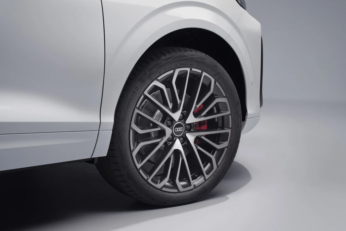 Biała karoseria samochodu oraz zbliżenie na nowoczesną, wieloramienną felgę aluminiową z logo Audi pośrodku, wokół której widoczna jest opona i czerwony zacisk hamulcowy.