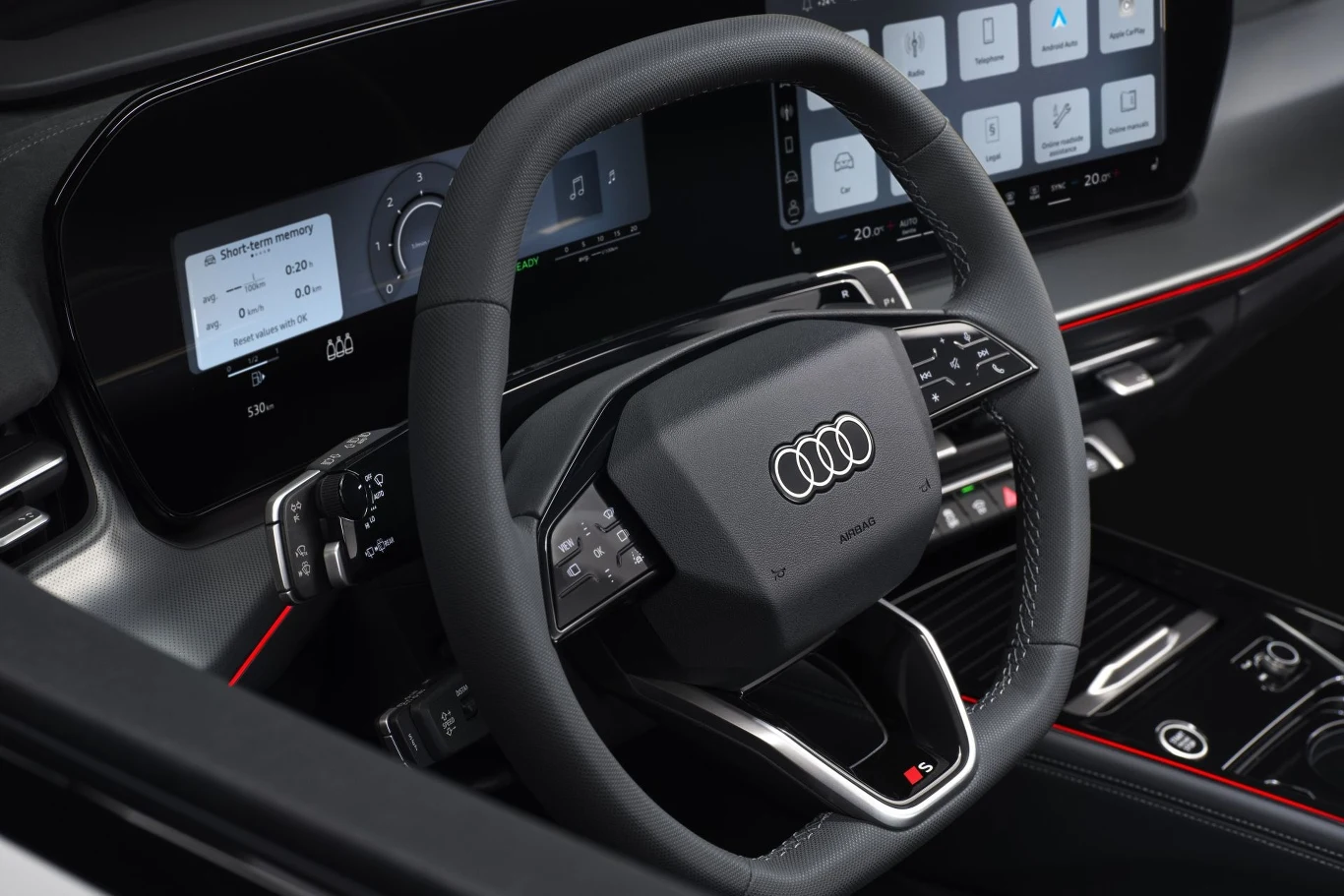 Kierownica samochodu marki Audi z nowoczesnym cyfrowym kokpitem, dużymi ekranami dotykowymi i eleganckimi detalami w czarnym oraz czerwonym kolorze.
