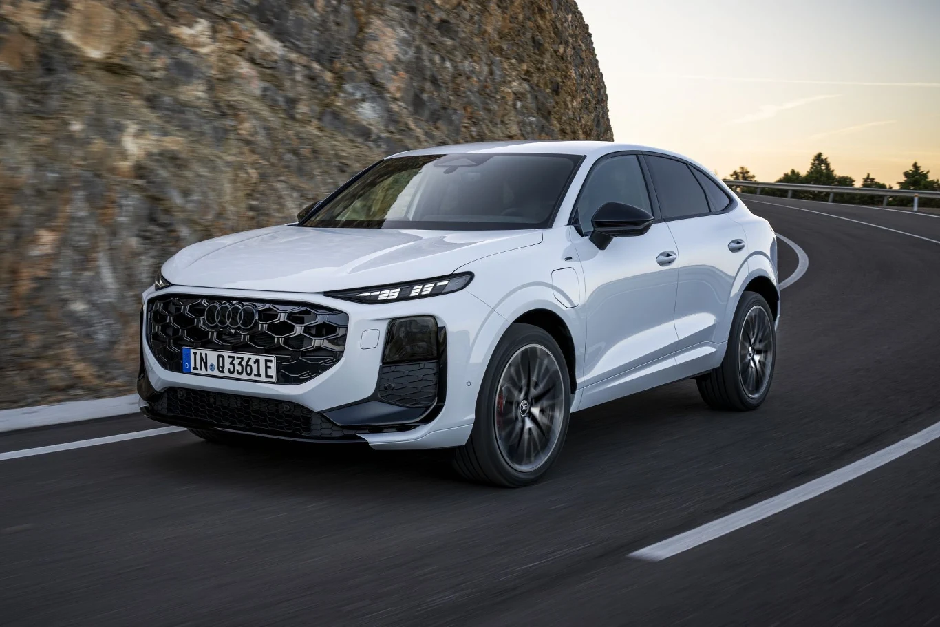 Biały samochód typu SUV marki Audi jedzie po krętej, asfaltowej drodze przy skalistej skarpie, w tle widoczny fragment nieba i pojedyncze drzewa.