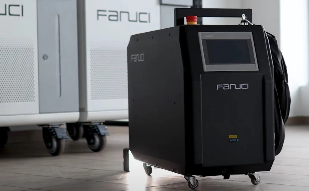 Przenośna spawarka laserowa Fanuci Czarna przemysłowa maszyna marki Fanuci na kółkach z panelem dotykowym i przewodami umieszczona na jasnej podłodze, w tle widoczne inne elementy urządzenia.