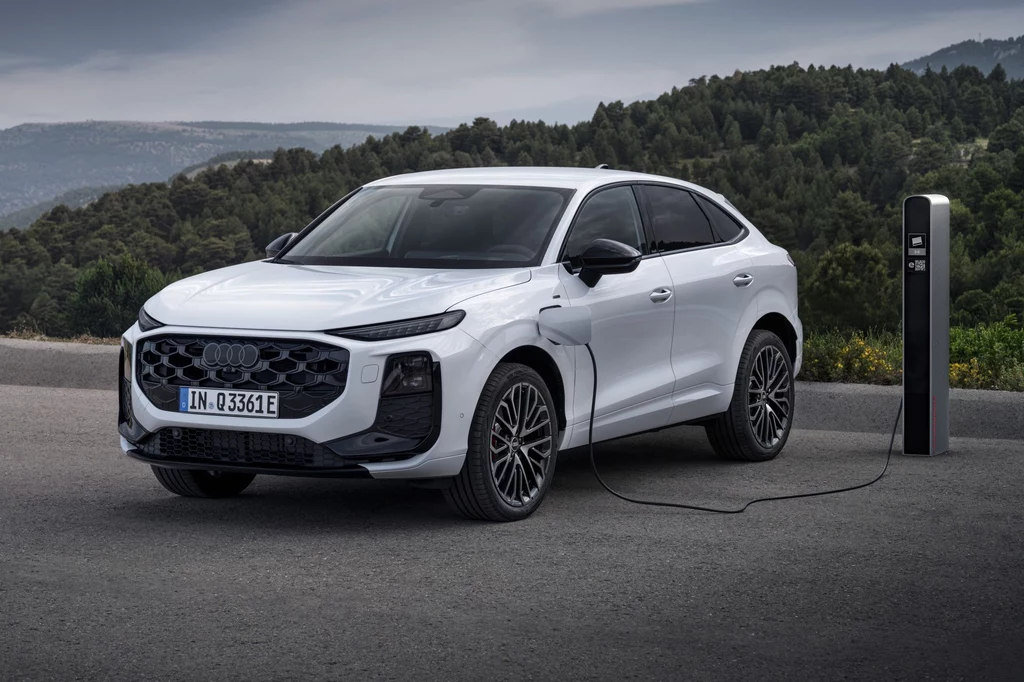 Audi Q3 Sportback dostępne jest również z hybrydą plug-in. Dzięki akumulatorowi o pojemności netto 19,7 kWh samochód może przejechać do 118 km na prądzie. Biały samochód marki Audi podłączony do stacji ładowania na leśnej trasie, w tle las i wzgórza.