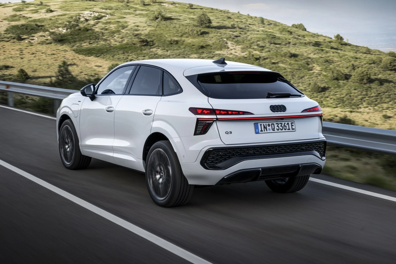 Biały samochód marki Audi Q3 jedzie po asfaltowej drodze w otoczeniu zielonych wzgórz, widoczne są czarne felgi oraz niemieckie tablice rejestracyjne.
