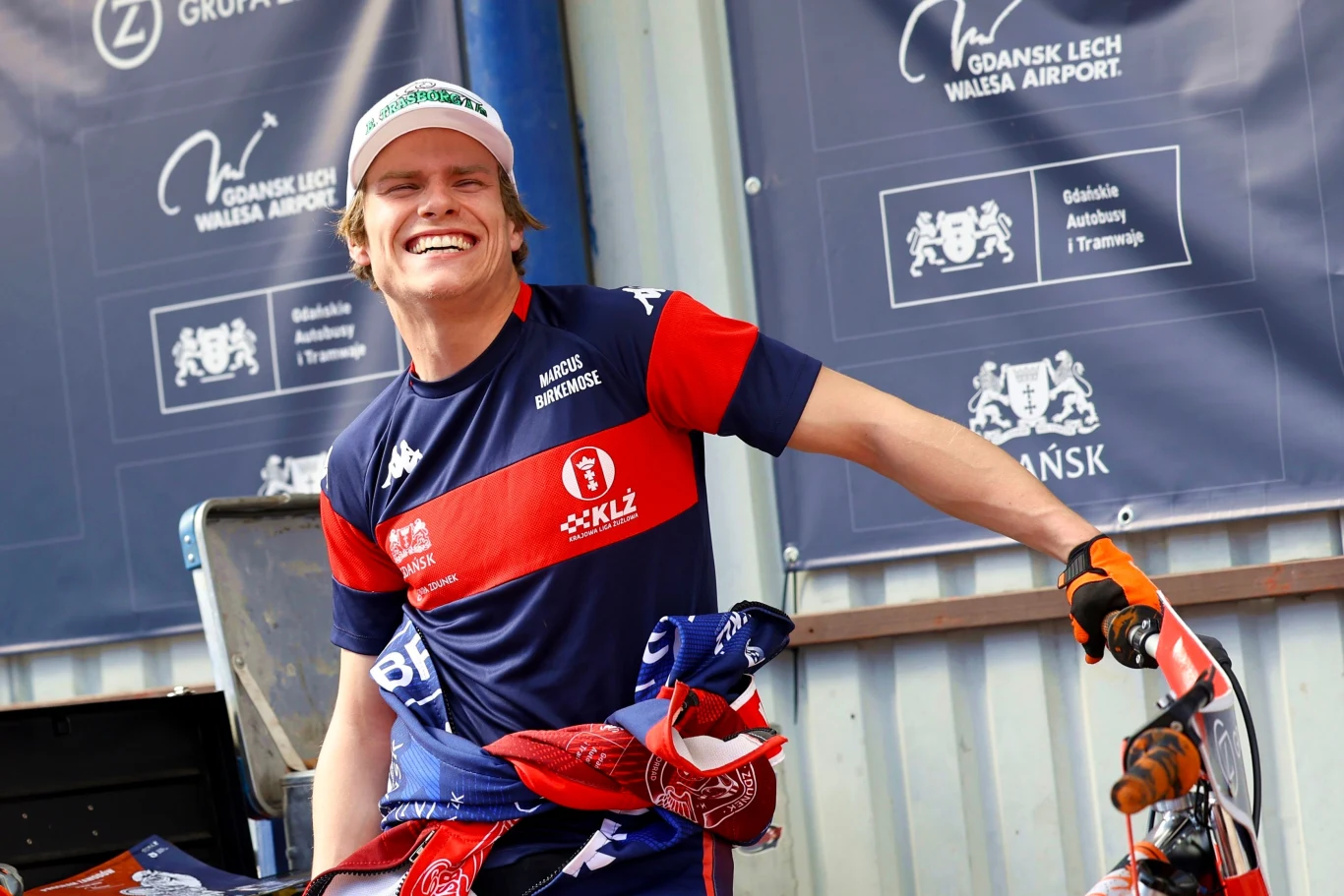 Zawodnik żużlowy uśmiechający się szeroko, ubrany w sportowy strój i czapkę z daszkiem, stoi obok motocykla w otoczeniu reklam i logotypów sponsorów imprezy.