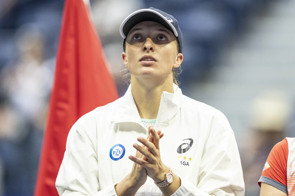 Iga Świątek wygrała US Open w 2022 roku Młoda kobieta ubrana w biały sportowy strój i czapkę z daszkiem, z poważnym i skupionym wyrazem twarzy, trzyma złożone dłonie przed sobą; w tle widoczna czerwona flaga oraz sylwetki innych osób.
