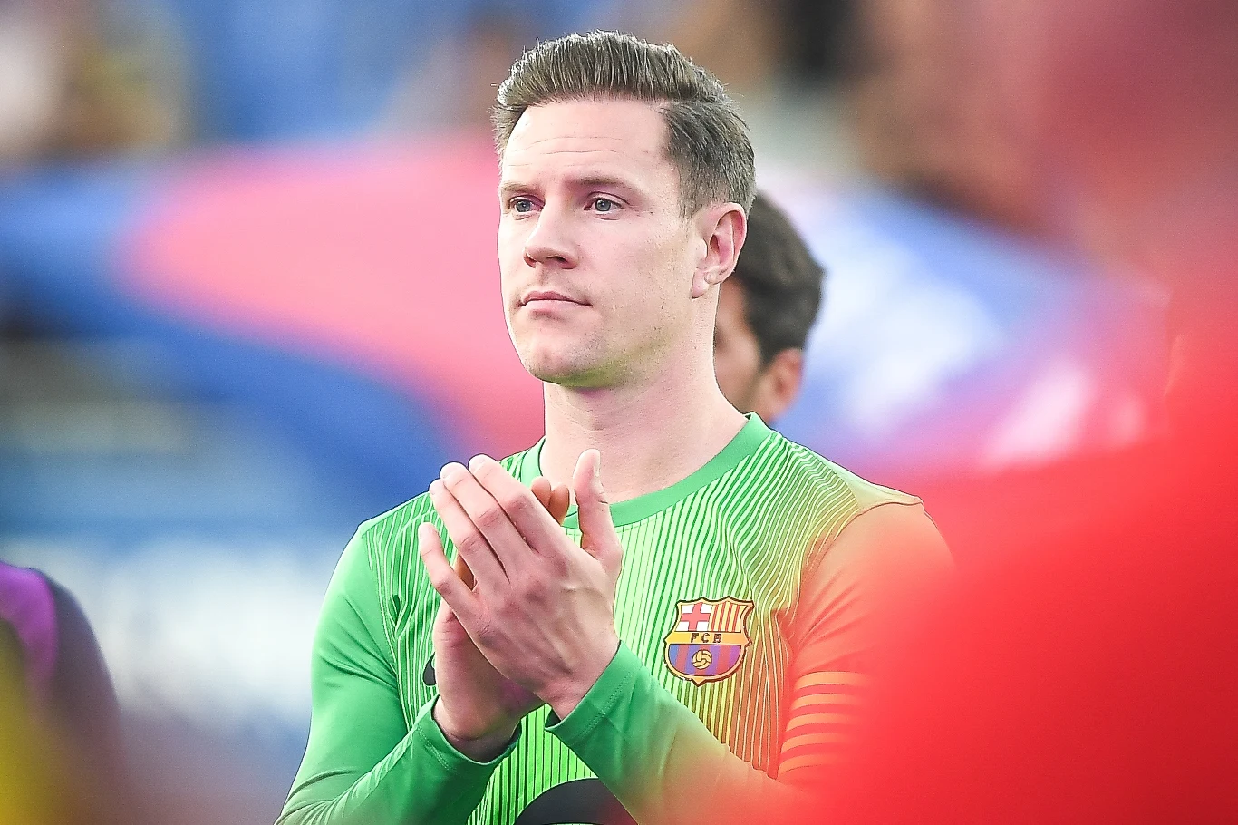 Marc-Andre ter Stegen Mężczyzna w zielonej koszulce piłkarskiej FC Barcelona stoi na tle rozmytego stadionu i klaszcze, jego wyraz twarzy jest skupiony.