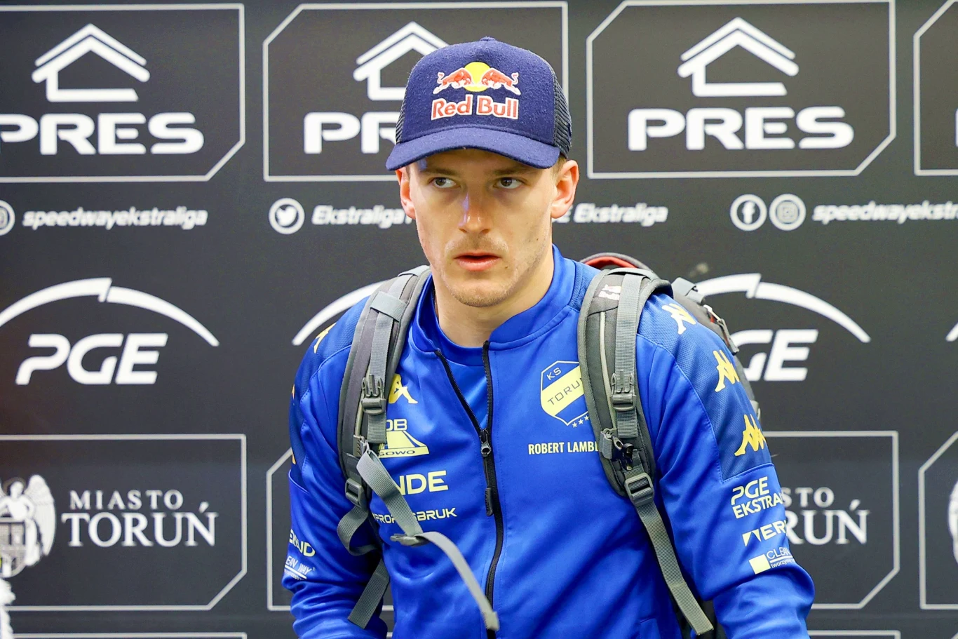 Mężczyzna w niebieskim stroju sportowym z logo zespołu i sponsorów, z plecakiem na plecach oraz czapką z daszkiem z logotypem Red Bull, stoi na tle tablicy z nazwami Miasto Toruń, PRES, PGE oraz logo Ekstraligi.