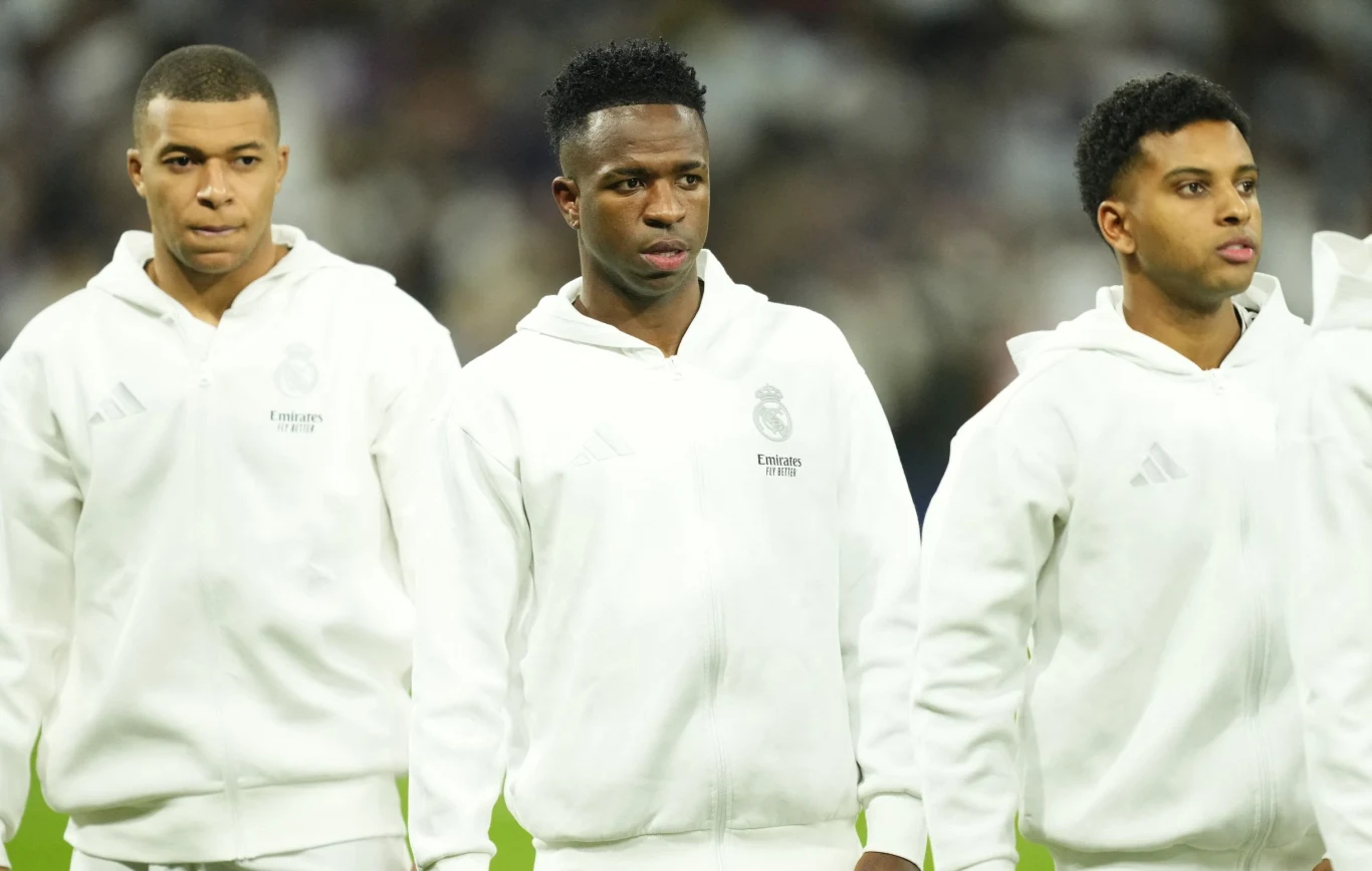 Kylian Mbappe, Vinicius Junior i Rodrygo Trzech młodych sportowców w białych dresach klubowych stoi obok siebie na murawie stadionu, patrząc przed siebie, w tle widoczna rozmyta publiczność.