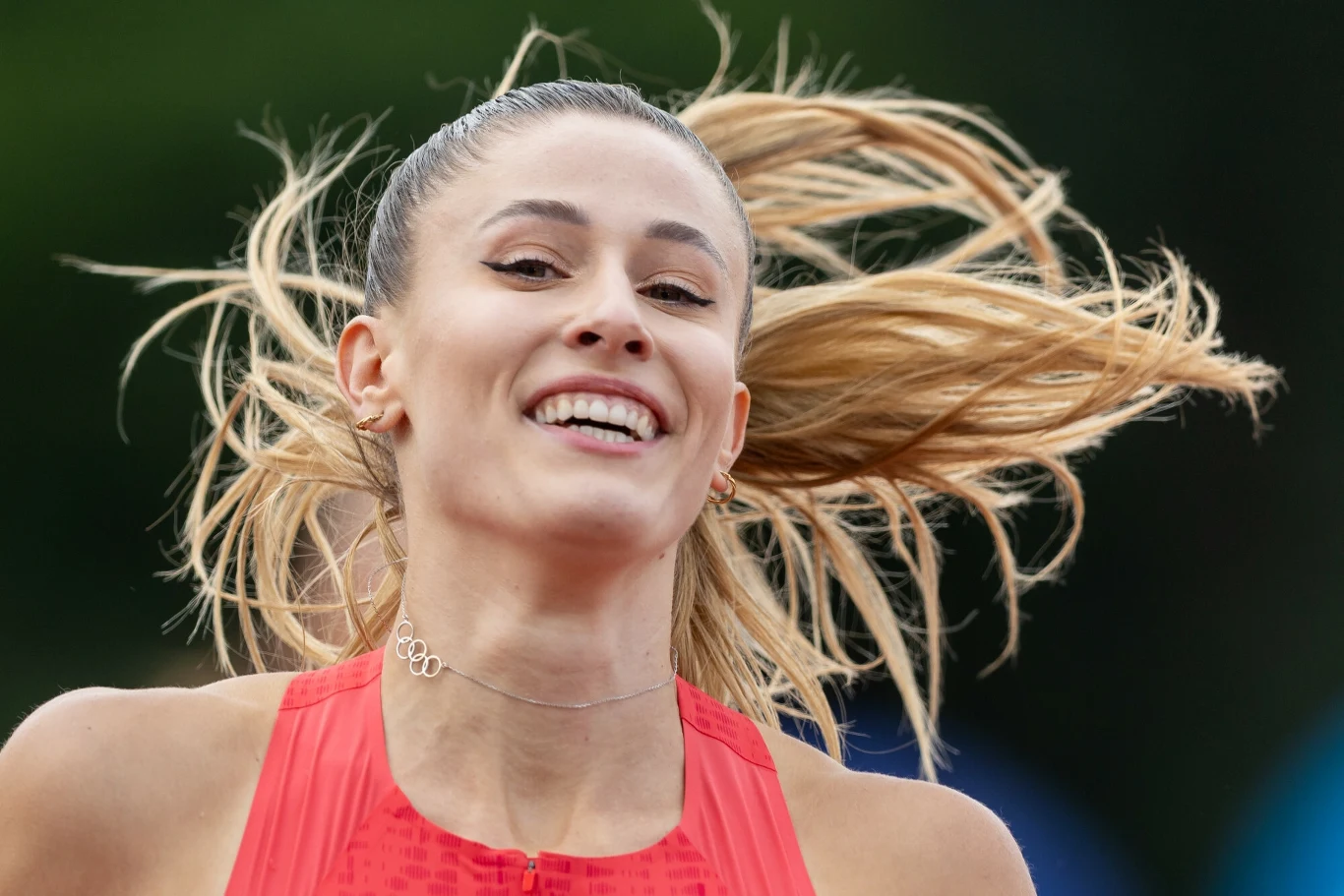 Uśmiechnięta młoda kobieta z długimi, rozwianymi blond włosami, ubrana w sportowy strój na tle rozmytego zielonego tła, chwyta dynamiczny moment podczas aktywności sportowej.