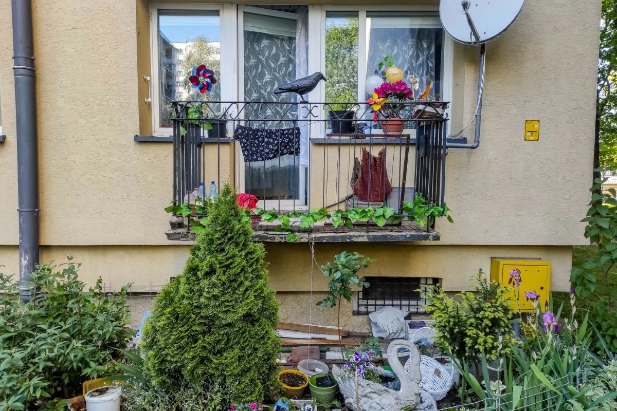Mały balkon z barierką udekorowany kolorowymi wiatraczkami, donicami z kwiatami i sztucznym ptakiem, poniżej gęsto obsadzony ogródek z krzewami, wieloma doniczkami oraz dekoracyjną figurką łabędzia i żaby.