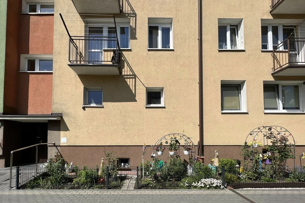 Nasadzenia w ogródku pod blokiem nie mogą naruszać przepisów o ochronie przyrody Blok mieszkalny z jasną elewacją, dwa balkony z metalowymi barierkami, zadbany ogródek ozdobny z kwiatami i dekoracjami ogrodowymi, ścieżka brukowa prowadząca wzdłuż budynku.