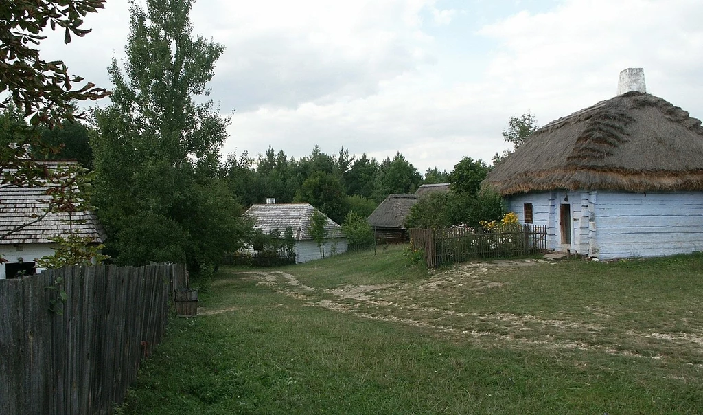 Muzeum Wsi Kieleckiej Stare, wiejskie zabudowania z charakterystycznymi strzechami na dachach, otoczone zielenią i drewnianym płotem, ścieżka prowadzi między domami przez zadbane podwórze, w tle drzewa i krzewy.