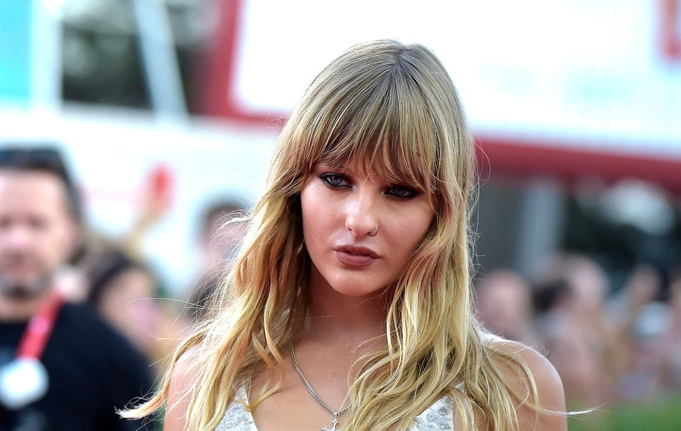 Victoria De Angelis jest basistką grupy Maneskin Młoda kobieta z długimi blond włosami i prostą grzywką patrząca w kierunku aparatu, w tle rozmyci ludzie i elementy architektury miejskiej.