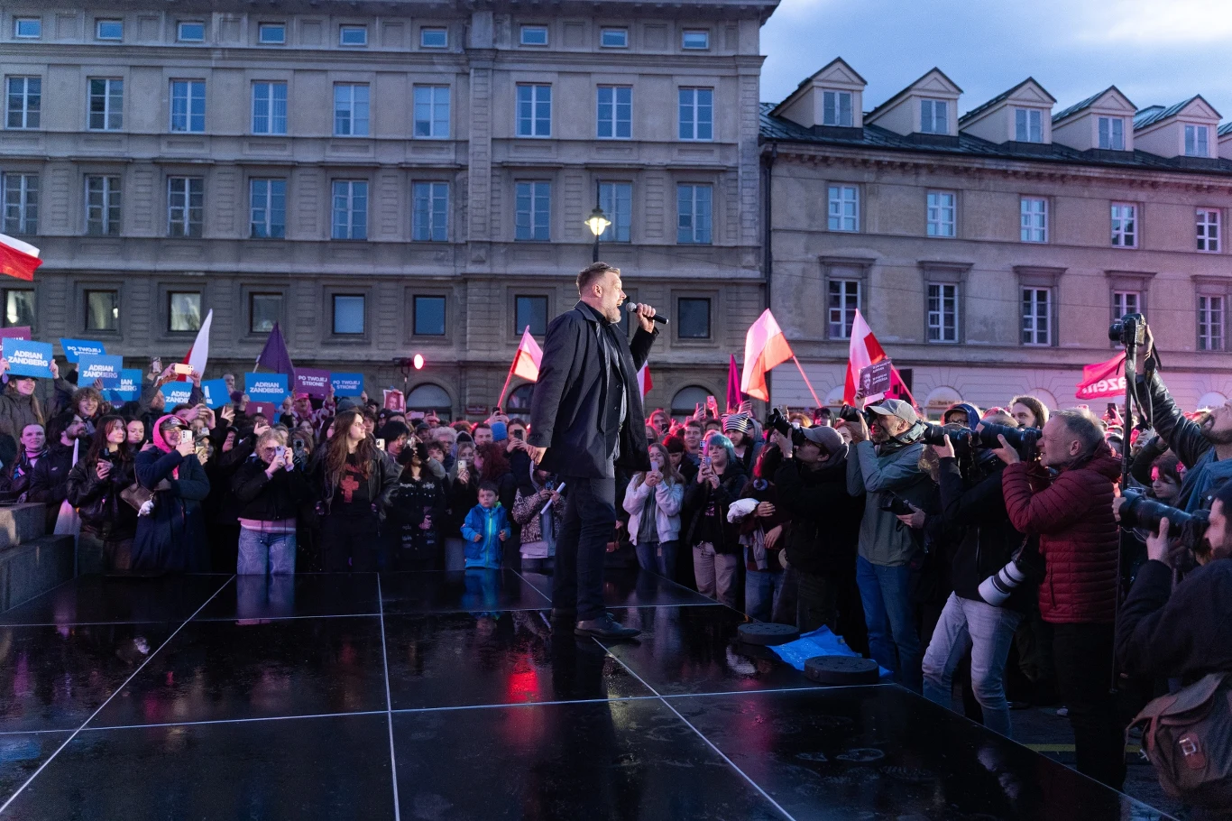 Tłum ludzi zgromadzony na otwartej przestrzeni w centrum miasta podczas wieczornego wydarzenia, na scenie mężczyzna przemawiający do zebranych, wokół liczne flagi, transparenty oraz fotografowie dokumentujący przebieg spotkania.