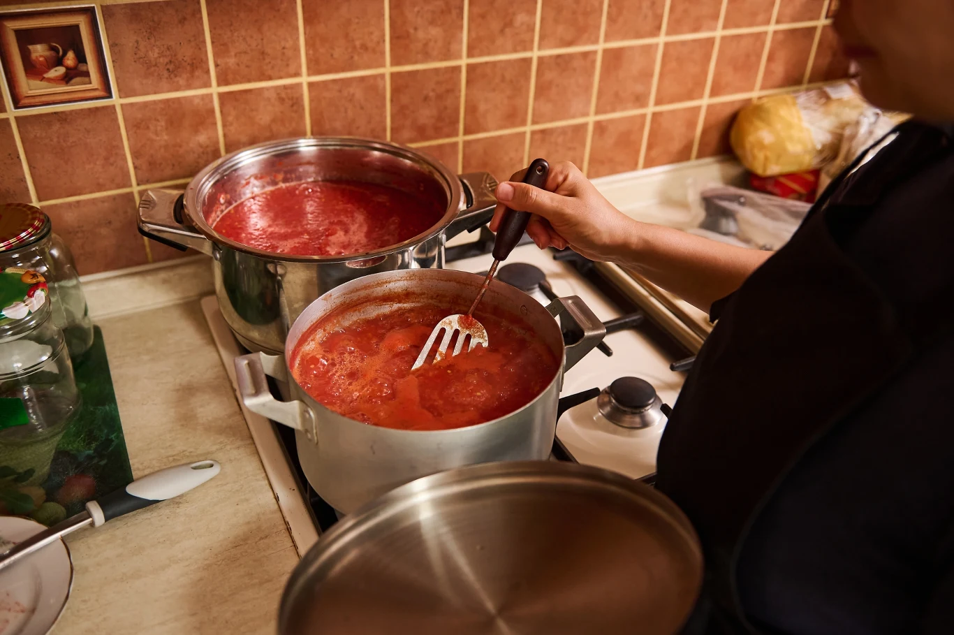 Kobieta miesza gotujący się sos pomidorowy w garnku na kuchence, obok stoi drugi garnek z podobną zawartością, całość w kuchennym otoczeniu z widocznym chlebem na blacie i pojemnikiem z warzywami.