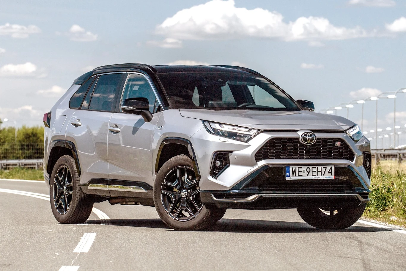 Od 1 stycznia Polacy jeszcze mocniej pokochają hybrydy plug-in? Powinny przyciągnąć głównie firmy Srebrny SUV Toyota RAV4 Hybrid zaparkowany na pustej drodze, w tle widoczna zielona roślinność oraz błękitne niebo z nielicznymi chmurami.