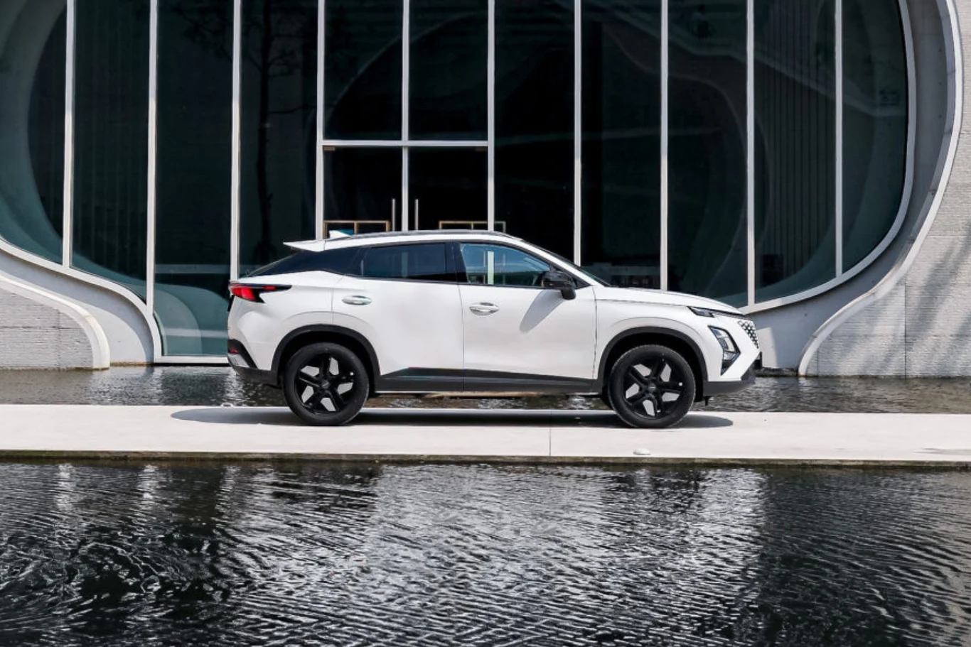 Biały samochód osobowy typu SUV stoi zaparkowany na jasnym chodniku nad brzegiem wody, w tle widoczna fasada nowoczesnego budynku ze szklanymi drzwiami i oknami o zaokrąglonych kształtach.