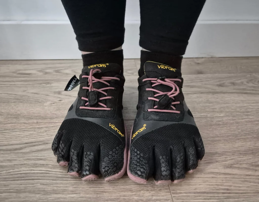 Buty typu barefoot Czarne buty typu barefoot z oddzielnymi palcami marki Vibram nałożone na stopy, czarne legginsy, jasna drewniana podłoga.