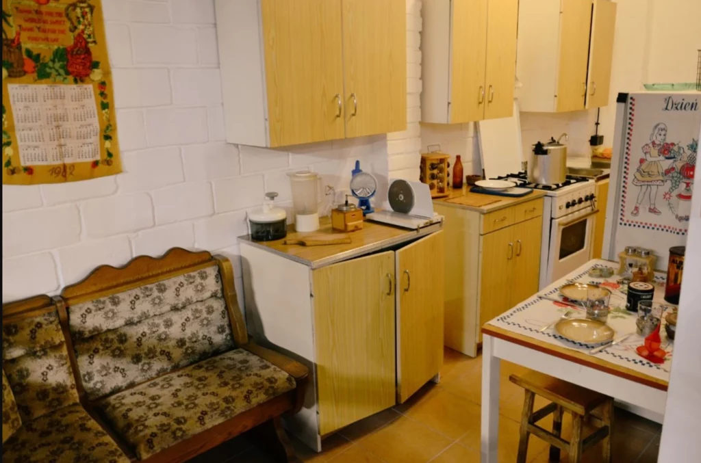 Tak często wyglądała kuchnia w bloku. To w niej mamy i babcie wymyślały wszystkie "patenty", które znamy do dziś Kuchnia urządzona w stylu vintage z drewnianymi szafkami, tapicerowaną kanapą, piecem gazowym oraz stołem z przygotowanymi produktami spożywczymi, po bokach widoczne kuchenne akcesoria i ozdobna ścierka na ścianie.