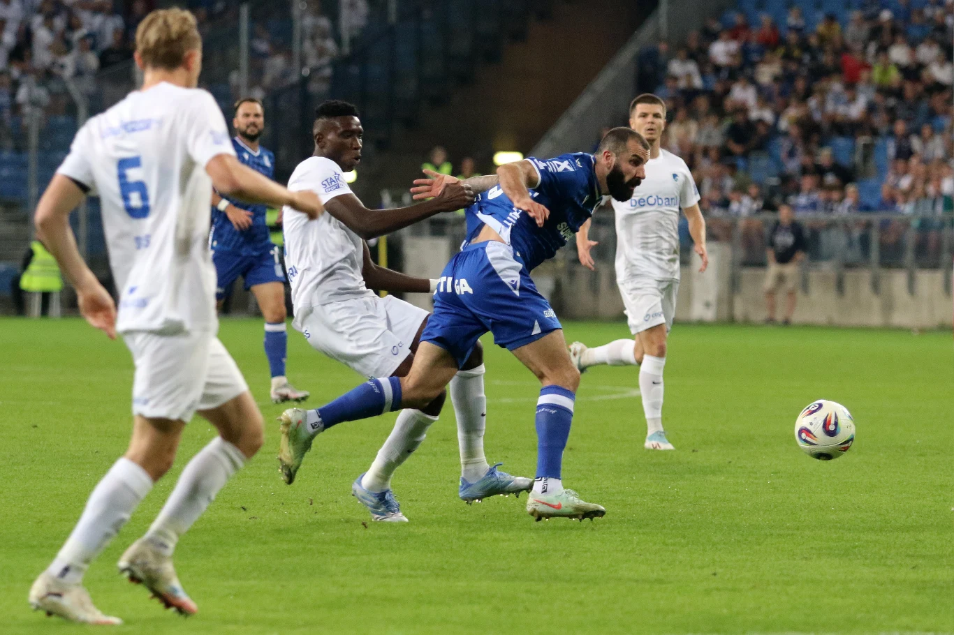 Zawodnicy dwóch drużyn piłkarskich walczą o piłkę na stadionie podczas meczu, wokół nich znajduje się wielu kibiców na trybunach.