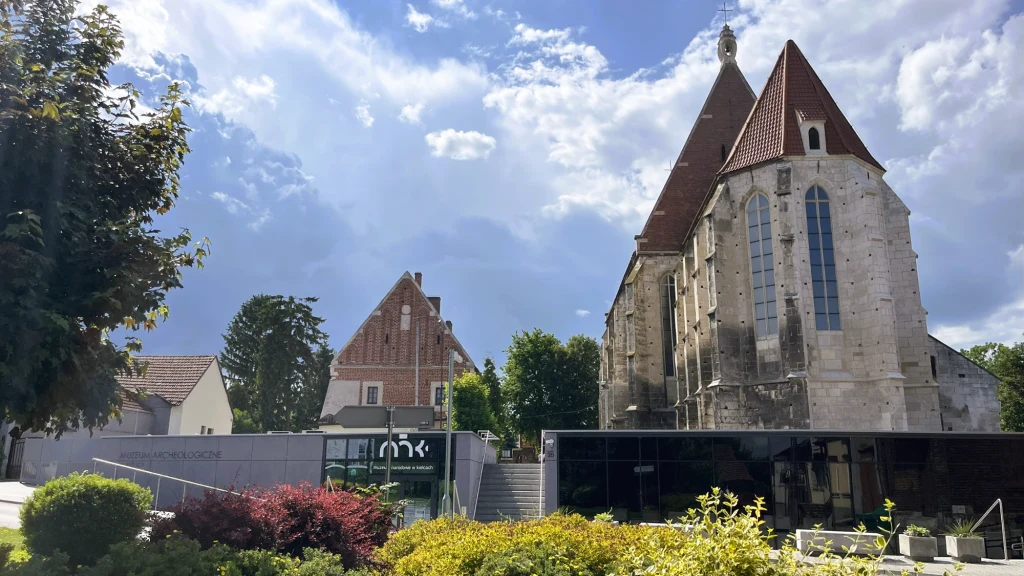 Zwiedzenie Muzeum Archeologicznego i Bazyliki pw. Narodzenia NMP to obowiązkowy punkt programu podczas wizyty w Wiślicy Dominująca gotycka budowla z charakterystycznym stromym dachem i kamienną fasadą, nowoczesny, przeszklony budynek w dolnej części kadru otoczony zielenią, obok widać kolejne historyczne budynki, całość oświetlona światłem dziennym z dynamicznym niebem ...