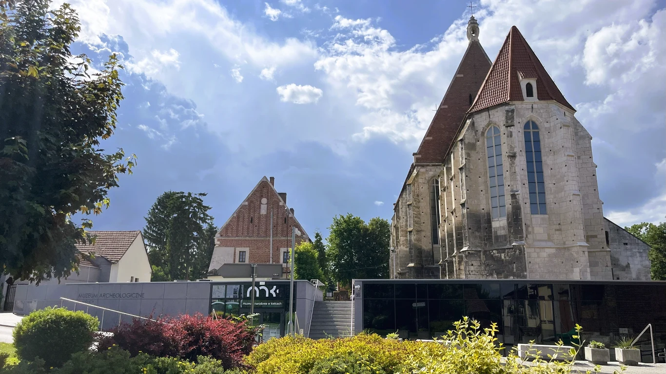 Dominująca gotycka budowla z charakterystycznym stromym dachem i kamienną fasadą, nowoczesny, przeszklony budynek w dolnej części kadru otoczony zielenią, obok widać kolejne historyczne budynki, całość oświetlona światłem dziennym z dynamicznym niebem ...