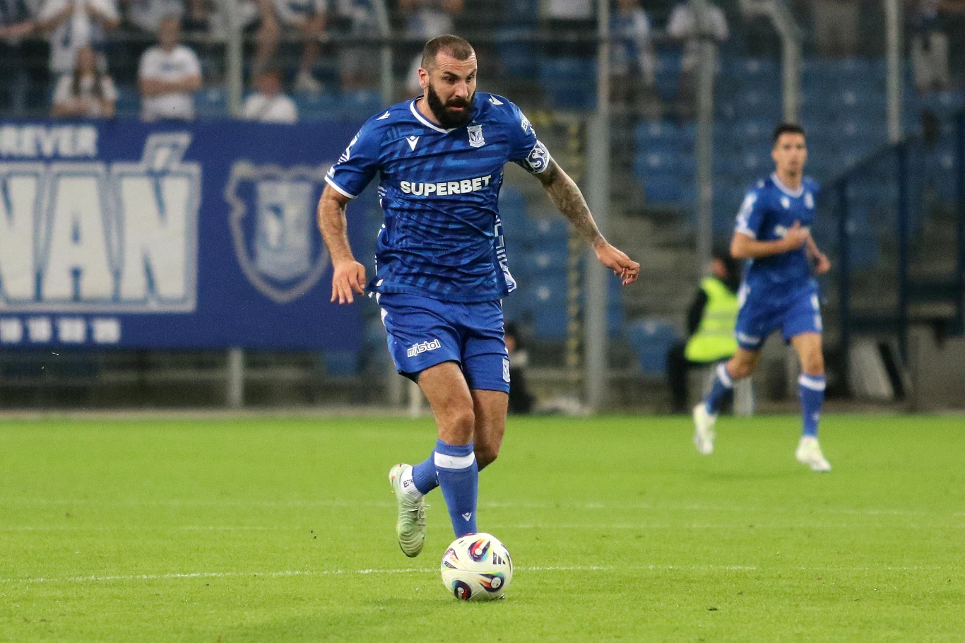 Piłkarz w niebieskim stroju prowadzi piłkę podczas meczu piłkarskiego na stadionie, w tle widoczni inni zawodnicy oraz fragment trybun z kibicami.
