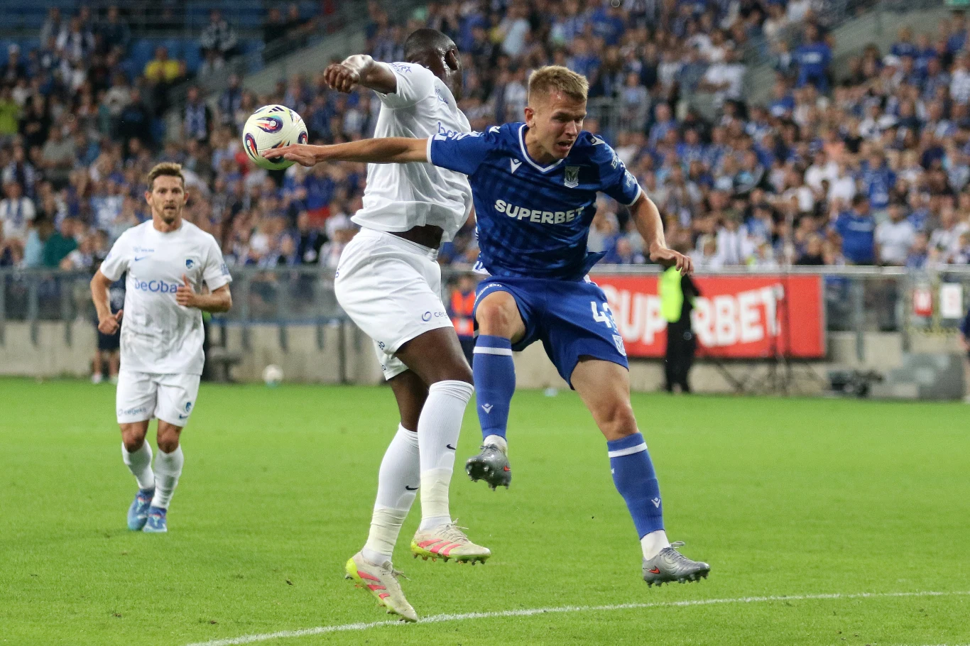 Dwóch piłkarzy walczy o piłkę podczas meczu piłki nożnej na stadionie, w tle widać tłum kibiców na trybunach.