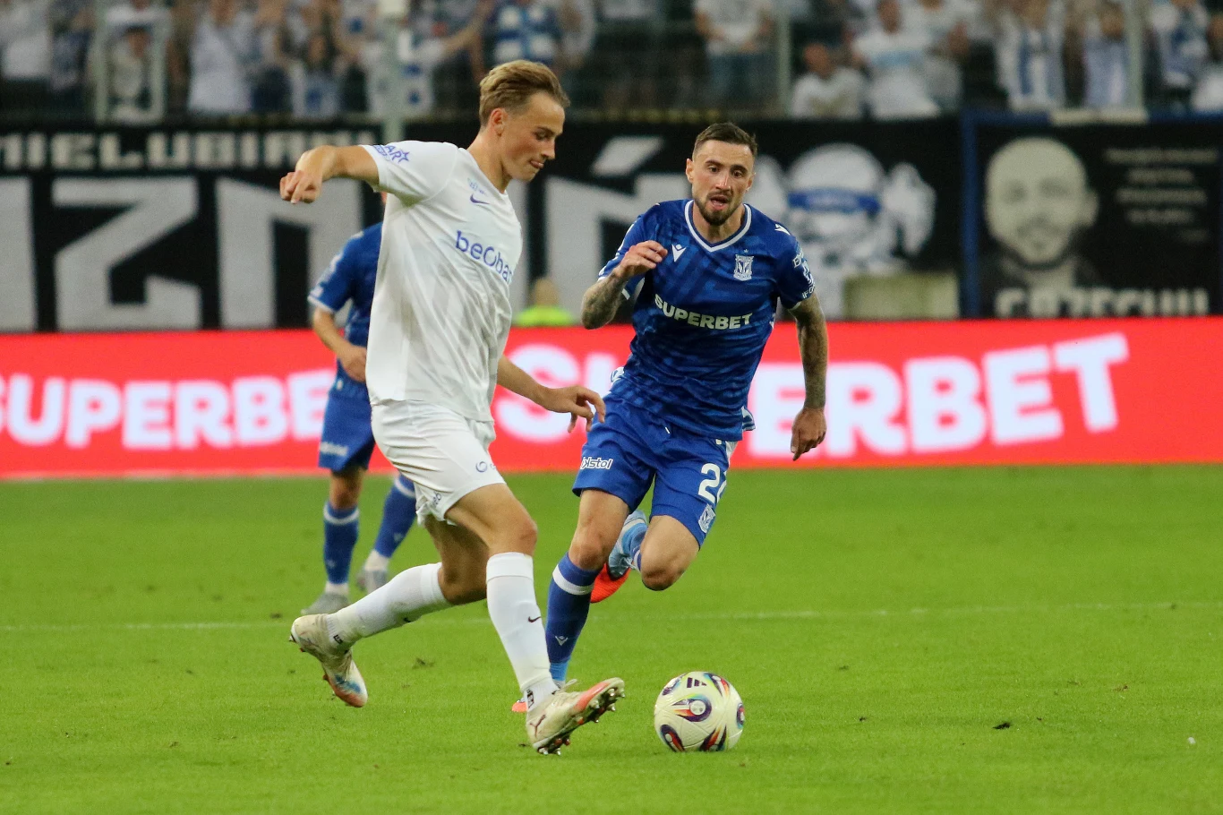 Filip Jagiełło, strzelec jedynego gola dla Lecha Poznań w meczu z Genk, w starciu z rywalem Dwóch piłkarzy w trakcie meczu, jeden ubrany w biały strój prowadzi piłkę, a zawodnik w niebieskim stroju stara się go dogonić, w tle widać rozmytych kibiców oraz reklamy na bandach stadionu.