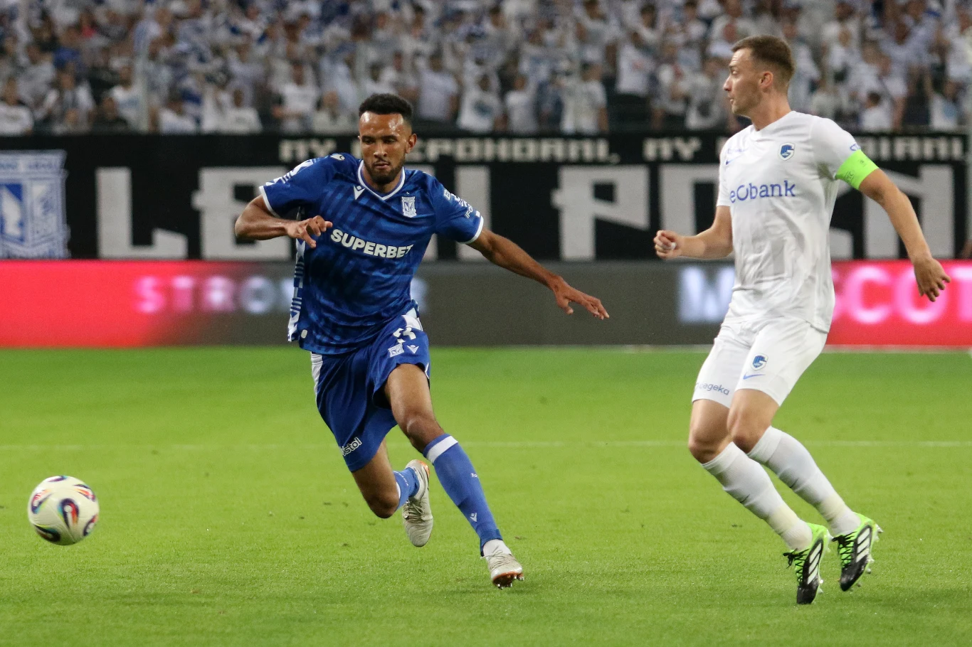 Dwóch piłkarzy w trakcie dynamicznej akcji na boisku, jeden ubrany w niebieski strój biegnie za piłką, drugi w białej koszulce obserwuje sytuację, w tle rozmyty tłum kibiców.