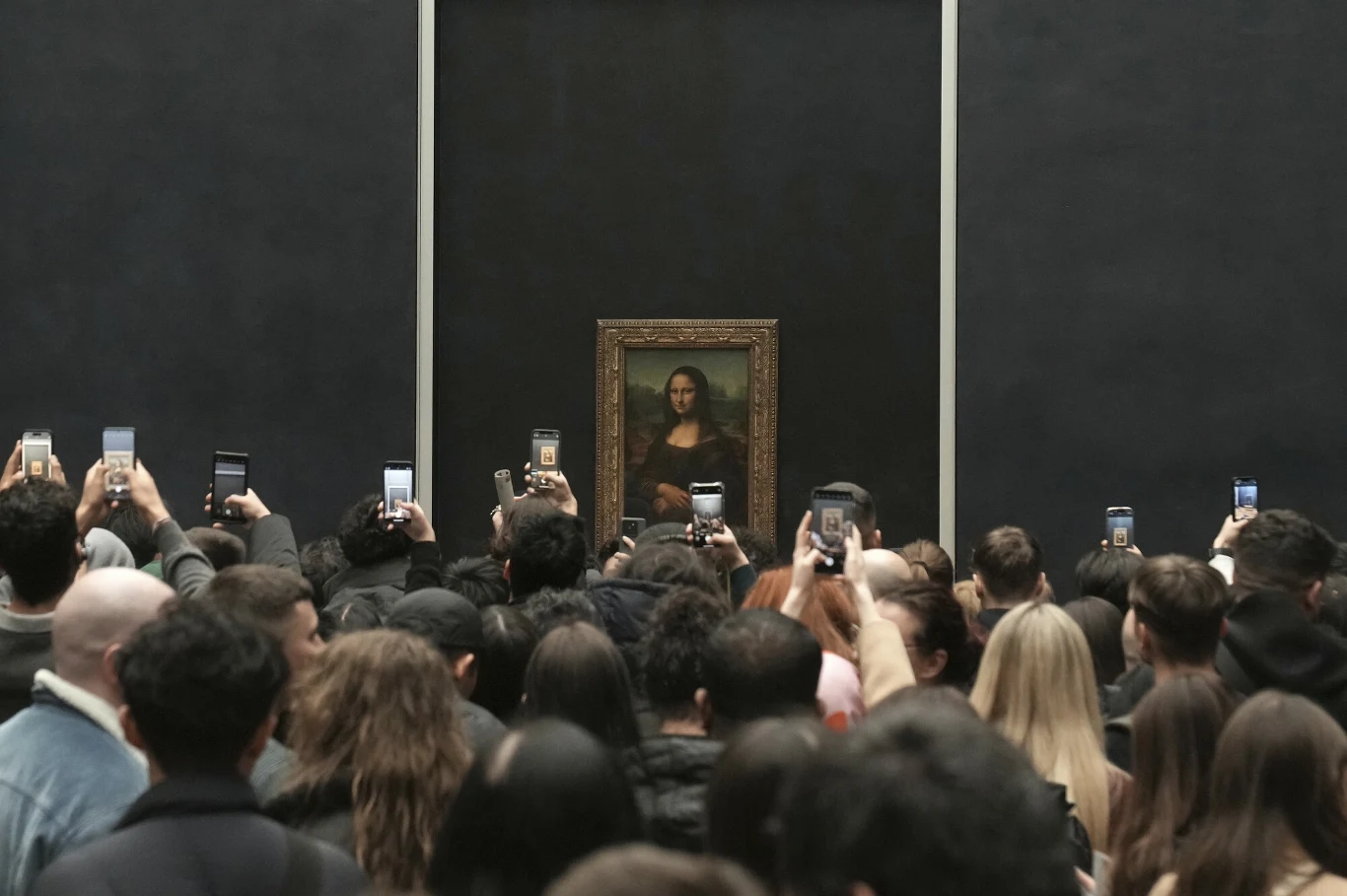 Luwr czeka wielki remont. Co dalej ze słynnym dziełem Leonarda da Vinci? Tłum ludzi skupiony przed obrazem Mona Lisa, większość zwiedzających kieruje telefony w stronę dzieła, wykonując zdjęcia lub nagrywając filmy, na pierwszym planie widoczna gęsto zgromadzona publiczność.