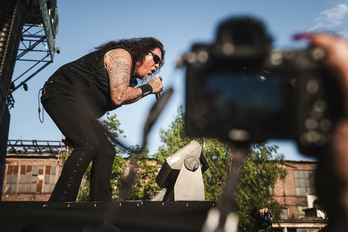 Chuck Billy (Testament) w akcji Wokalista z długimi, ciemnymi włosami i tatuażami na ramionach wykonuje utwór na scenie podczas koncertu plenerowego, w tle widoczne są stare zabudowania oraz wyciągnięta w stronę sceny kamera lub aparat.