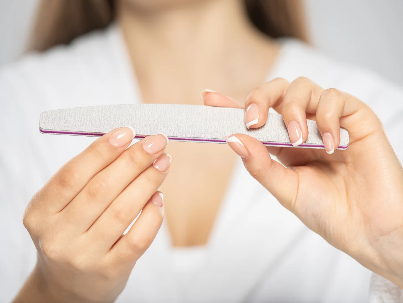 Sposoby na trwały lakier na paznokciach Dłonie kobiety trzymające pilnik do paznokci, z zadbanymi, starannie wypiłowanymi paznokciami, w tle rozmyta biała odzież.